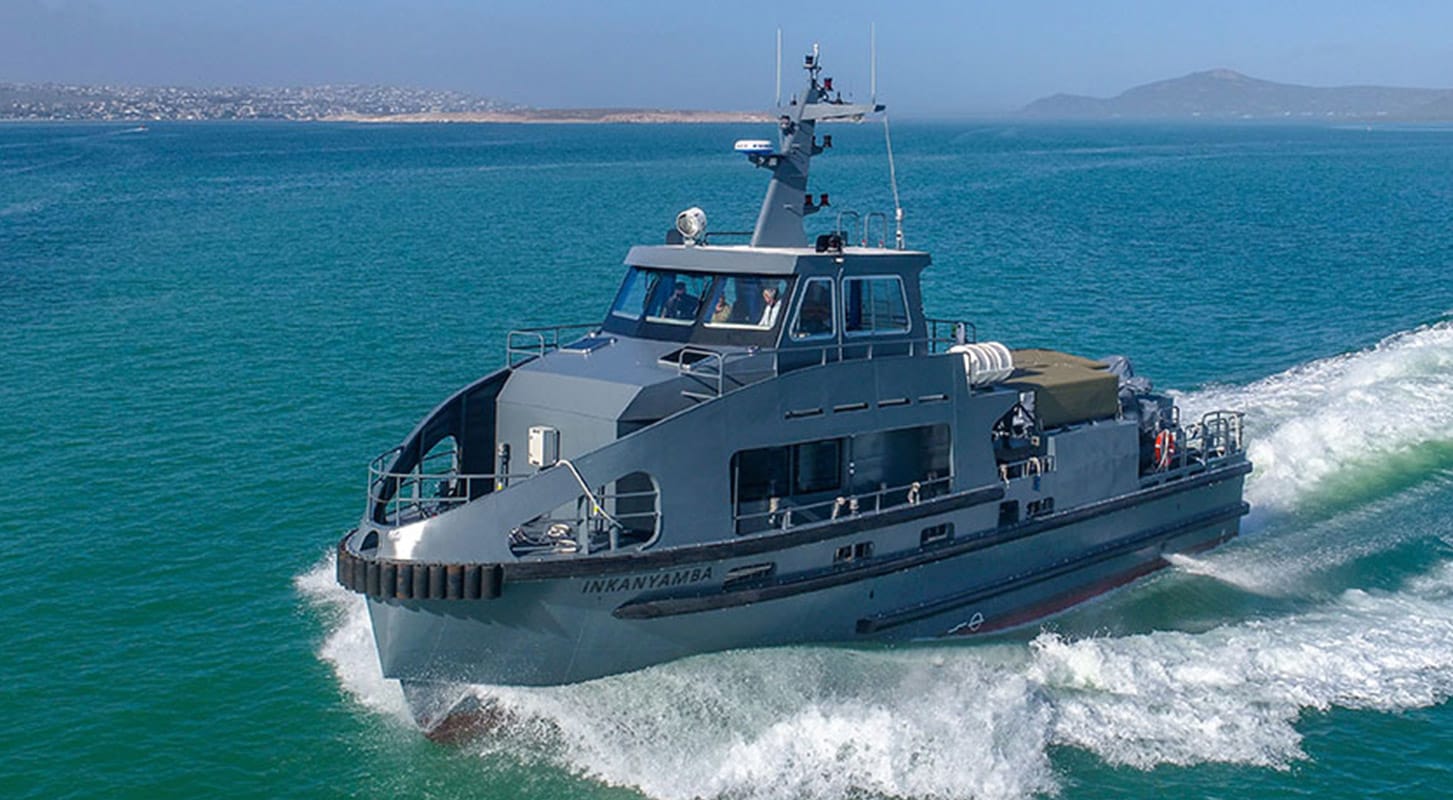 A gray patrol boat named "Inkan'yamba" speeds through greenish-blue water near a coastline with hills in the background.